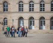 Eine Gruppe junger Leute schlendert gemeinsam vor dem historischen Gebäude der Hochschule Coburg, das durch seine Rundbogenfenster und zahlreichen Türen charakterisiert ist. Lässig gekleidet unterhalten sie sich angeregt unter dem sonnigen Himmel.