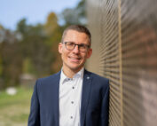 Ein Mann in blauem Anzug und weißem Hemd lehnt draußen an einer reflektierenden Wand der Hochschule Coburg. Er lächelt unter seiner Brille und steht vor einem verschwommenen Hintergrund aus Bäumen und Grün unter einem klaren Himmel.