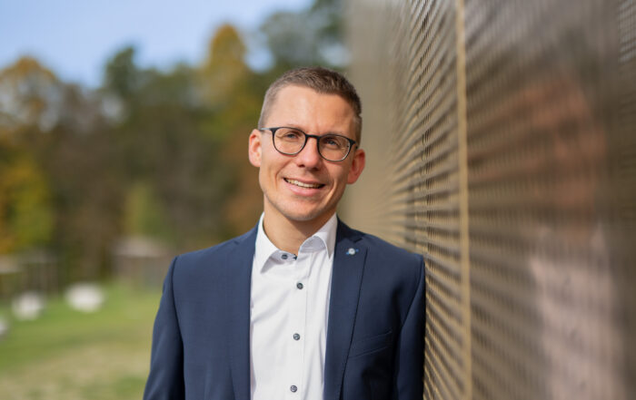 Ein Mann in blauem Anzug und weißem Hemd lehnt draußen an einer reflektierenden Wand der Hochschule Coburg. Er lächelt unter seiner Brille und steht vor einem verschwommenen Hintergrund aus Bäumen und Grün unter einem klaren Himmel.