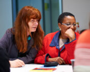 Zwei Menschen, einer mit langen roten Haaren, der andere mit kurzen Haaren, sitzen an einem Tisch in der Hochschule Coburg. Sie wirken konzentriert, die rechte Person stützt ihr Kinn auf die Hand. Papiere und Notizbücher liegen verstreut auf dem Tisch.