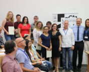 Eine Gruppe von Menschen steht in einem Hörsaal der Hochschule Coburg zusammen, einige halten Zertifikate in den Händen. Sie lächeln, einige sitzen im Vordergrund. Im Hintergrund ist ein Banner zu sehen.