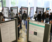 Eine Gruppe von Menschen bei einer Posterpräsentation der Hochschule Coburg in einem geräumigen Raum mit großen Fenstern. Die Teilnehmer betrachten verschiedene wissenschaftliche Poster, die auf Ständen ausgestellt sind, und diskutieren darüber. Der Boden ist gefliest und der Raum ist von natürlichem Licht durchflutet.