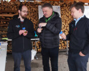 Drei Männer stehen zusammen an der Hochschule Coburg und untersuchen im Freien kleine Flaschen. Hinter ihnen sind Holzscheite gestapelt. Zwei der Männer tragen dunkle Jacken mit Logos. Im Hintergrund sind Tische mit weiteren Flaschen und Informationstafeln zu sehen, die Innovation und Forschung hervorheben.