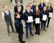 Eine Gruppe von Menschen steht im Innenbereich der Hochschule Coburg, einige halten Zertifikate in den Händen. Sie tragen Jacken und lächeln in die Kamera, um die Essenz dieses offiziellen akademischen Ereignisses einzufangen.