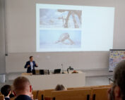 In einem Hörsaal der Hochschule Coburg präsentiert ein Mann im Anzug Bilder von Science-Fiction-Robotern auf einer Projektionsfläche. Die Zuhörer sitzen, und seitlich davon sind auf Staffeleien lebendige Illustrationen zu sehen.