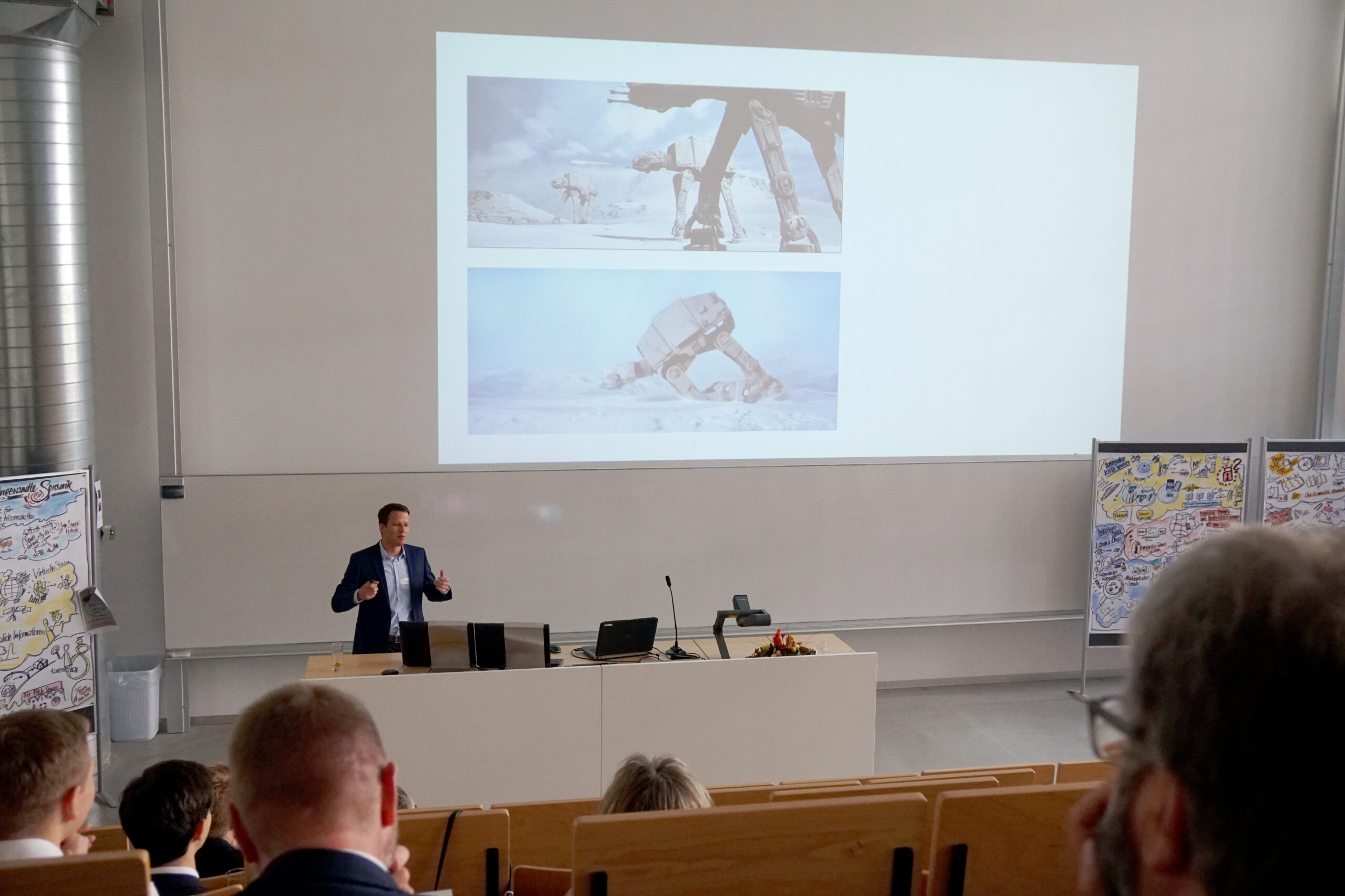 In einem Hörsaal der Hochschule Coburg präsentiert ein Mann im Anzug Bilder von Science-Fiction-Robotern auf einer Projektionsfläche. Die Zuhörer sitzen, und seitlich davon sind auf Staffeleien lebendige Illustrationen zu sehen.