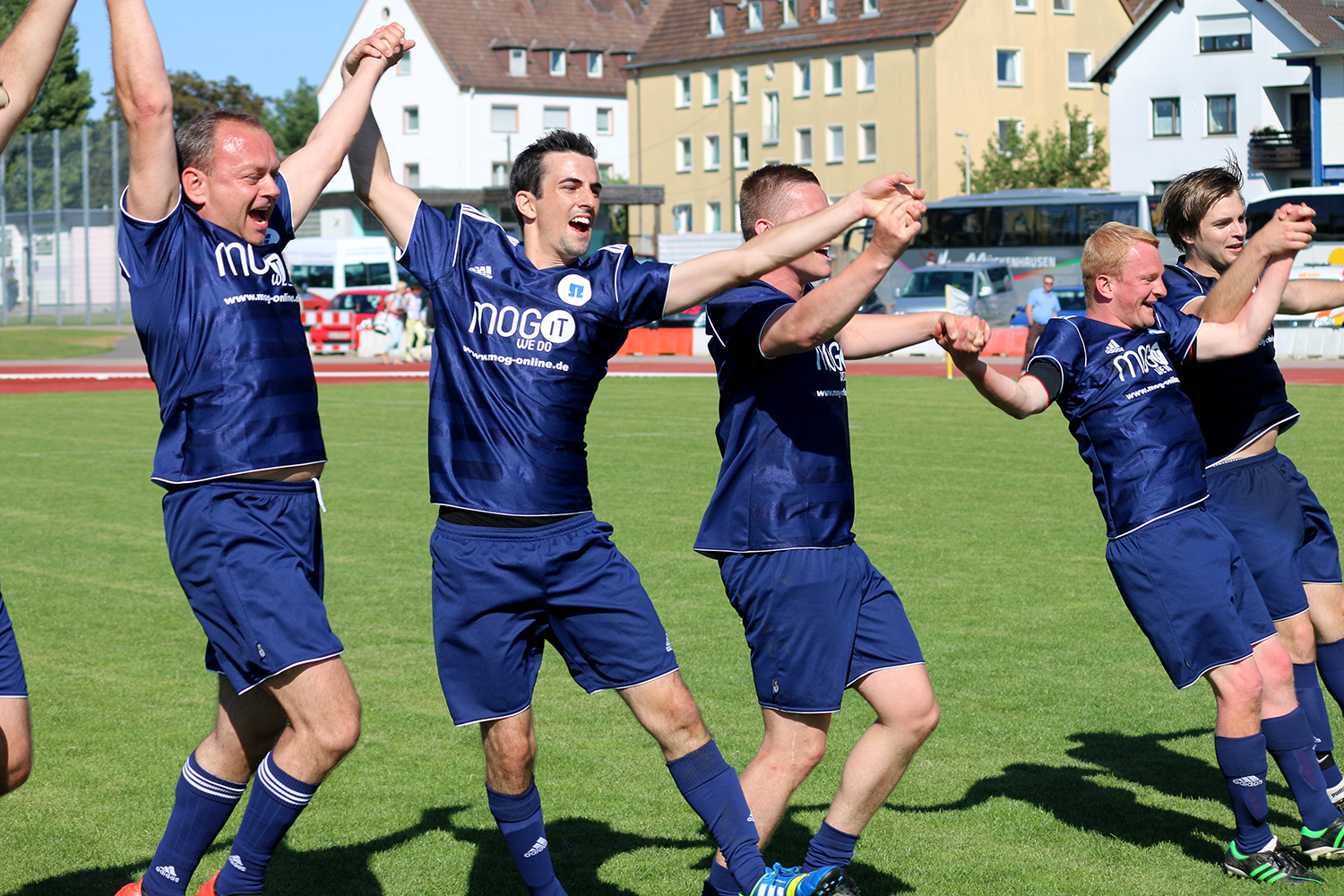 Eine Gruppe jubelnder Fußballspieler der Hochschule Coburg in blauen Trikots feiert auf einem Feld, hält sich an den Händen und reckt siegreich die Arme in die Höhe. Im Hintergrund sind eine Laufbahn, einige Gebäude und Bäume unter einem klaren blauen Himmel zu sehen.