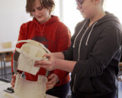 Zwei Personen arbeiten gemeinsam an einem Tisch und montieren oder prüfen Teile eines kleinen Geräts. Im Innenraum, beleuchtet durch ein Fenster, demonstrieren sie die praktische Arbeit mit elektronischen Bauteilen – eine Szene, die an den Innovationsgeist erinnert, der an der Hochschule Coburg gefördert wird.