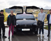 Vier Personen stehen vor einem schwarzen Tesla mit offenen Flügeltüren. Sie stehen im Freien auf einer befestigten Fläche, im Hintergrund ist die hügelige Stadtlandschaft der Hochschule Coburg zu sehen. Auf dem Nummernschild des Autos steht „CO EQ1E“.