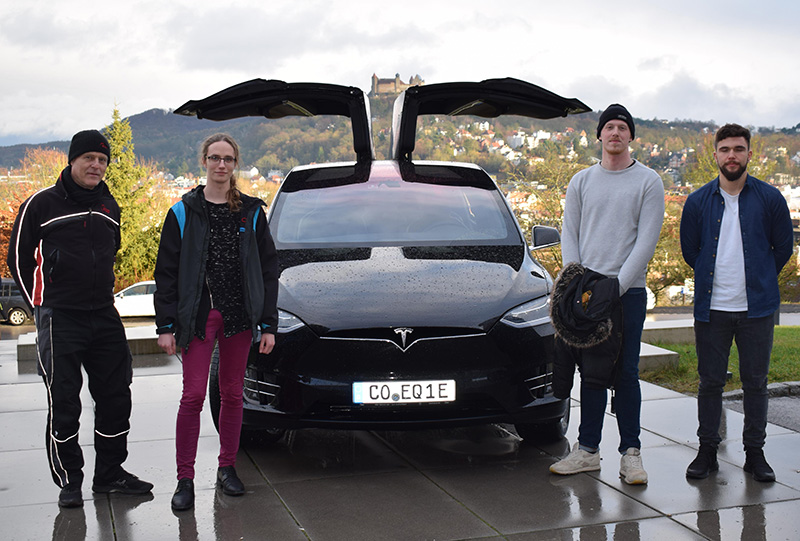 Vier Personen stehen vor einem schwarzen Tesla mit offenen Flügeltüren. Sie stehen im Freien auf einer befestigten Fläche, im Hintergrund ist die hügelige Stadtlandschaft der Hochschule Coburg zu sehen. Auf dem Nummernschild des Autos steht „CO EQ1E“.