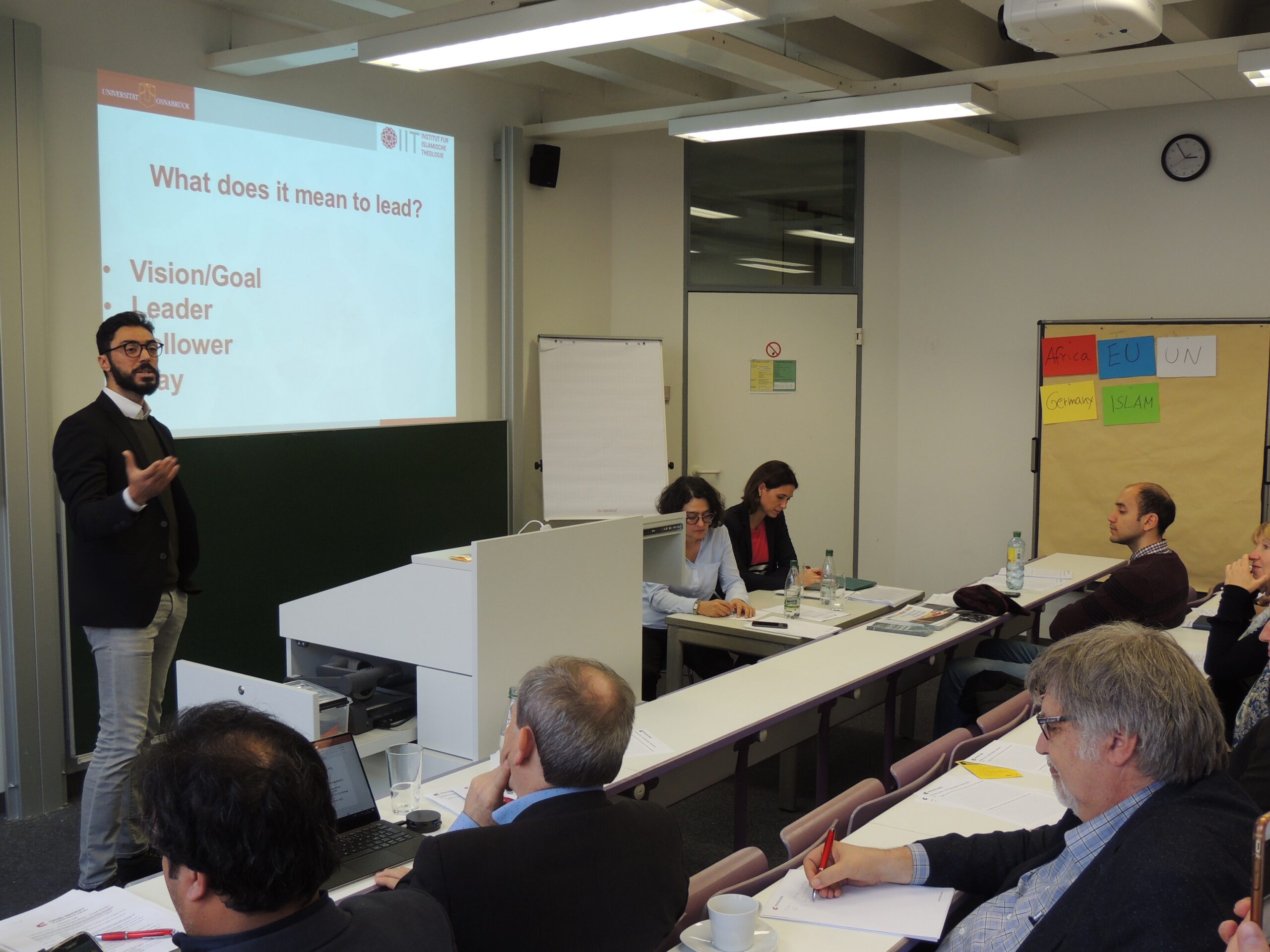 Ein Mann hält vor einer Gruppe an der Hochschule Coburg einen Vortrag. Er steht neben einem Bildschirm, auf dem eine Folie mit dem Titel „Was bedeutet es, zu führen?“ und Aufzählungspunkten zu sehen ist. Die Teilnehmer sitzen an Tischen, machen sich Notizen und hören aufmerksam zu.