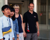 Vier Menschen stehen vor einem Gebäudeeingang der Hochschule Coburg. Links zwei junge Erwachsene in Freizeitkleidung, rechts neben der Glastür mit der Aufschrift „Hochschule Coburg“ stehen eine Frau und ein Mann.