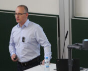 Eine Person mit Brille und hellblauem Hemd hält in einem Hörsaal der Hochschule Coburg einen Vortrag. Sie steht mit einem Laptop und einer Flasche Wasser neben einem Podium, während hinter ihr teilweise eine Projektionsfläche zu sehen ist.