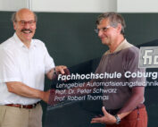 Zwei Männer lächeln und halten ein transparentes Schild mit dem Text „Hochschule Coburg“, dem Namen „Fachhochschule Coburg“ und Angaben zum „Lehrgebiet Automatisierungstechnik“ mit den Namen zweier Professoren hoch. Sie stehen vor einer Tafel.