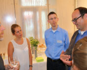 Vier Personen versammeln sich in einem Raum zu einer lebhaften Diskussion. Ein Mann mit Brille gestikuliert lebhaft, während er spricht, und zieht damit die Aufmerksamkeit einer Frau in einem weißen Oberteil auf sich, die aufmerksam zuhört. Zwei weitere Personen beteiligen sich mit engagierten Mienen, und im Hintergrund sind Erfrischungen zu sehen, die an Treffen der Hochschule Coburg erinnern.