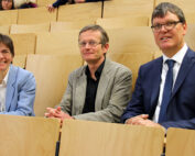 Drei Personen sitzen in einem Hörsaal der Hochschule Coburg. Die Person links trägt eine hellblaue Jacke, die mittlere eine graue und die rechte einen marineblauen Anzug. Sie blicken nach vorne und sitzen bequem auf hellen Holzstühlen.