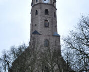 Ein hoher, historischer Uhrenturm aus Stein ragt an der Hochschule Coburg in den Himmel, umgeben von kahlen Bäumen. Der Turm verfügt über ein Spitzdach mit kleinen Dachgauben und einem auf der Vorderseite sichtbaren Zifferblatt.