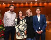 Vier Personen stehen zusammen in einem gut beleuchteten Raum der Hochschule Coburg, umgeben von Holzwänden und einer kreisförmigen Anordnung. In formeller Kleidung wirken sie selbstsicher und professionell, während auf der linken Seite elegante Blumenarrangements zu sehen sind.