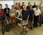 Eine Gruppe von sechzehn Männern, einige in Freizeitkleidung, steht drinnen um ein großes mechanisches Gerät auf einem Metallrahmen herum. Die Szene spielt in einer Werkstatt oder einem Labor der Hochschule Coburg.