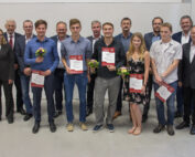 Eine Gruppe von Menschen posiert im Innenbereich der Hochschule Coburg. Vier Personen in der ersten Reihe halten Zertifikate und kleine Blumensträuße, während andere in formeller Kleidung hinter ihnen stehen. Die Szene lässt auf eine Anerkennungs- oder Preisverleihung an dieser angesehenen Institution schließen.