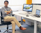 Eine Person sitzt im Schneidersitz auf einem Bürostuhl an einem Schreibtisch, auf dem ein Desktop-Computer und ein Laptop stehen. Auf dem Bildschirm ist ein Auto zu sehen. Sie trägt ein kariertes Hemd, beige Hosen und braune Schuhe und befindet sich im Büro der Hochschule Coburg mit seinen weißen Wänden und Jalousien.