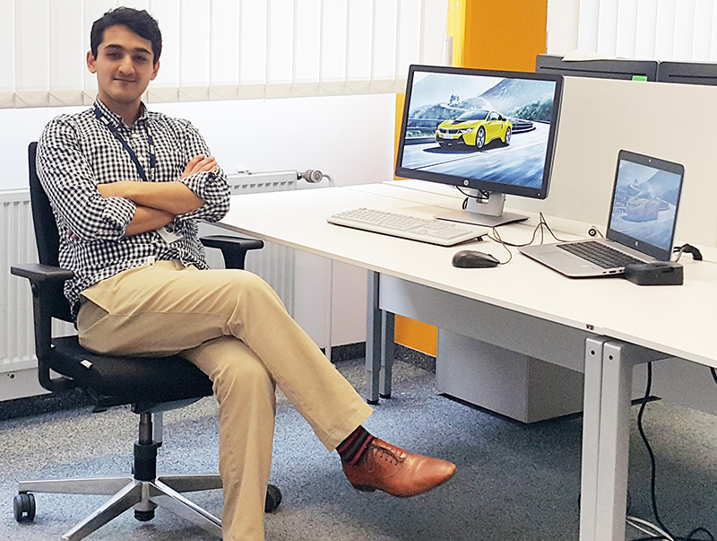 Eine Person sitzt im Schneidersitz auf einem Bürostuhl an einem Schreibtisch, auf dem ein Desktop-Computer und ein Laptop stehen. Auf dem Bildschirm ist ein Auto zu sehen. Sie trägt ein kariertes Hemd, beige Hosen und braune Schuhe und befindet sich im Büro der Hochschule Coburg mit seinen weißen Wänden und Jalousien.