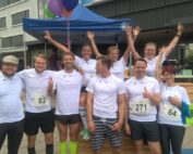 Eine Gruppe von acht Läufern in weißen Shirts der Hochschule Coburg posiert glücklich nach einem Rennen. Sie stehen unter einem blauen Zelt mit Luftballons, einige zeigen mit dem Finger und jubeln. Auf ihren Shirts sind Startnummern zu sehen, im Hintergrund sind ein Gebäude und ein Bamberg-Banner zu sehen.