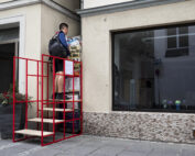 Eine Person steht auf einem roten, gestuften Präsentationsständer, der an einem Gebäude angebracht ist, das an das architektonische Flair der Hochschule Coburg erinnert, und hängt ein Plakat an eine Wand. Die Struktur ähnelt einer Treppe mit Flyer-Werbung. In der Nähe steht eine große Topfpflanze, während das Gebäude mit einem Glasfenster und einer beigefarbenen Fassade aufwartet.