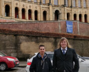 Zwei Personen stehen vor einem kunstvollen, historischen Gebäude mit Bogenfenstern und dekorativem Mauerwerk, das an die Architektur der Hochschule Coburg erinnert. Die Person links trägt eine dunkle Jacke, die Person rechts einen Anzug. Entlang der Straße stehen geparkte Autos.