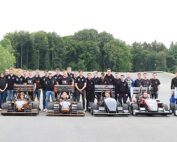 Eine große Gruppe von Menschen steht auf einer Straße hinter mehreren eleganten, farbenfrohen Rennwagen, die in einer Reihe aufgereiht sind. Im Hintergrund sind die üppigen Grünflächen und hohen Bäume der Hochschule Coburg zu sehen. Die einzelnen Personen tragen passende Hemden, was auf Teamgeist oder ein gemeinsames Ziel hindeutet.