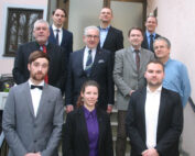 Eine Gruppe von zehn Personen, darunter Männer und Frauen, in formeller Kleidung, posiert für ein Foto auf den Stufen eines Gebäudes der Hochschule Coburg. Sie sind in zwei Reihen angeordnet, wobei einige elegant auf den Stufen stehen und andere auf dem Boden sitzen.