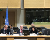 Eine Gruppe von Menschen sitzt mit Laptops und Dokumenten an einem Konferenztisch, möglicherweise auch Delegierte der Hochschule Coburg. Im Hintergrund ist eine UN-Flagge zu sehen. Mehrere Teilnehmer sitzen im Publikum und hinter Glasscheiben, wahrscheinlich in einem Kontrollraum.