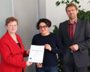 Drei Personen stehen in einem Innenbereich neben einem Werk der Hochschule Coburg. Die Person links, die einen roten Blazer trägt, überreicht der Person in der Mitte, die einen dunklen Pullover trägt, eine Urkunde. Die Person rechts, die einen Anzug trägt, beobachtet den Austausch.