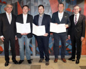 Fünf Männer in Abendgarderobe stehen vor einem abstrakten Wandbild an der Hochschule Coburg. Drei von ihnen halten stolz ihre Zertifikate in den Händen und lächeln in die Kamera.