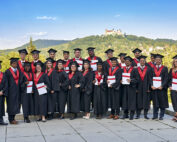 Eine Gruppe von Absolventen der Hochschule Coburg in schwarzen Talaren und Hüten posiert im Freien. Jeder hält ein Diplom in der Hand, während sie auf einer Steinplattform stehen und im Hintergrund einen malerischen Blick auf Hügel und eine Burg unter einem klaren blauen Himmel genießen.