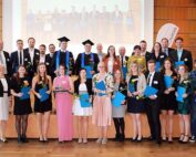 Eine Gruppe von Menschen posiert drinnen, einige tragen Abschlusskappen und Talare und halten stolz Diplome und Blumen in den Händen. Einige Personen sind elegant in Anzügen und Kleidern gekleidet. Hinter ihnen zeigt ein projiziertes Bild ihren denkwürdigen Tag an der Hochschule Coburg.