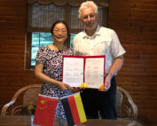 Eine Frau und ein Mann stehen in einem Raum der Hochschule Coburg und halten stolz ein Zertifikat in den Händen. Sie lächeln, vor ihnen steht ein Tisch mit kleinen chinesischen und deutschen Flaggen. Der Raum hat Holzwände und ein Fenster mit Jalousien.