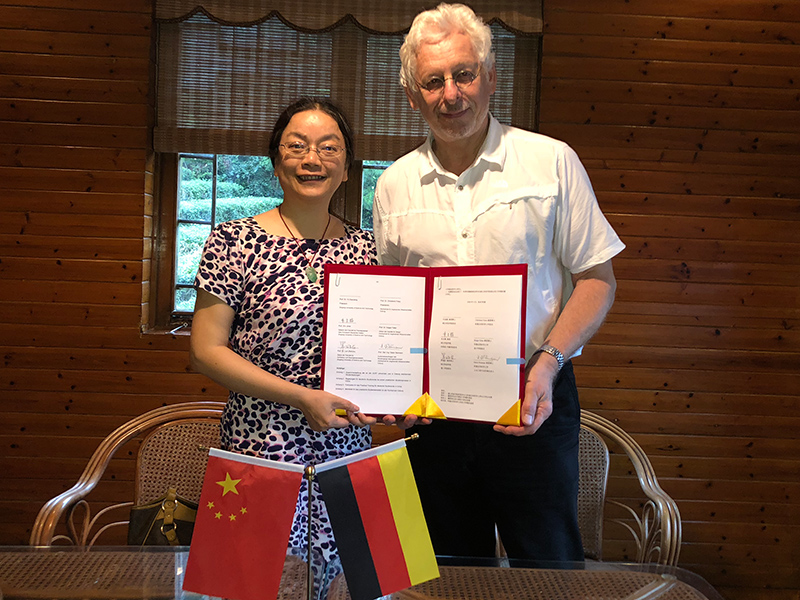Eine Frau und ein Mann stehen in einem Raum der Hochschule Coburg und halten stolz ein Zertifikat in den Händen. Sie lächeln, vor ihnen steht ein Tisch mit kleinen chinesischen und deutschen Flaggen. Der Raum hat Holzwände und ein Fenster mit Jalousien.