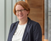 Eine Person mit kurzen roten Haaren und Brille lächelt. Sie trägt einen dunklen Blazer über einem weißen Hemd. Sie steht in einem Innenbereich der Hochschule Coburg, wo der unscharfe Hintergrund subtile Details hellgrauer und brauner Paneele offenbart.