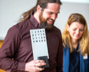 Ein Mann mit Bart und kastanienbraunem Hemd lächelt und hält einen Designpreis der Hochschule Coburg in den Händen. Neben ihm steht eine Frau mit blonden Haaren und blauem Hemd und teilt seine Freude. Sie befinden sich in einem Raum mit einer Präsentationsleinwand als Hintergrund.