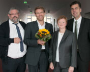 Vier Personen stehen in einem Flur der Hochschule Coburg. Drei Männer und eine Frau sind in Business-Kleidung gekleidet und strahlen eine formelle Atmosphäre aus. Ein Mann hält einen Blumenstrauß mit einer Sonnenblume in der Hand und alle lächeln herzlich.