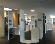 Der moderne Ausstellungsbereich der Hochschule Coburg besteht aus weißen Ausstellungsständen, die in einem geräumigen Flur angeordnet sind. Jeder Stand präsentiert Texte und Bilder zu Umweltthemen. Der Raum ist mit natürlichem und künstlichem Licht gut beleuchtet, während Pflanzen die Ecken schön schmücken.