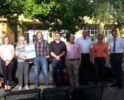 Eine Gruppe von acht Personen, leger gekleidet und lächelnd, steht vor einem gelben Gebäude mit einer Skulptur unter einem Baum an der Hochschule Coburg. Eine Person hält stolz eine Urkunde in den Händen und hält damit einen Moment der Leistung fest.