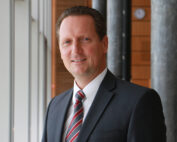 Eine Person in Anzug und gestreifter Krawatte steht im Innenbereich neben großen Fenstern und lächelt in die Kamera. Der Hintergrund, der an die moderne Ästhetik der Hochschule Coburg erinnert, zeigt architektonische Elemente mit Metall- und Holzdetails.