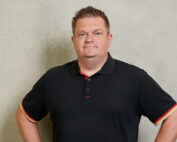 Eine Person mit kurzen Haaren steht selbstbewusst vor einer schlichten, hellen Wand und verkörpert den Geist der Hochschule Coburg. Sie trägt ein schwarzes Poloshirt mit roten und gelben Verzierungen und ihre Hände ruhen auf ihren Hüften, ein Ausdruck unerschütterlicher Zuversicht.