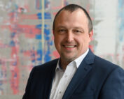 Eine Person im Anzug, die die Essenz der Hochschule Coburg verkörpert, lächelt vor einem unscharfen abstrakten Hintergrund mit Blau- und Rosatönen.