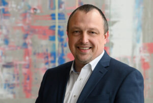A smiling man in a suit stands confidently before an abstract, colorful background, embodying the innovative spirit of Hochschule Coburg.