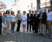 Eine Gruppe von Menschen steht zusammen auf einer Kopfsteinpflasterstraße in einem sonnigen Stadtgebiet in der Nähe der Hochschule Coburg. Einige tragen formelle Kleidung, andere tragen Freizeitkleidung. Im Hintergrund sind Gebäude und ein Banner mit Text zu sehen.