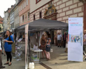 Auf einem lebhaften Straßenfest steht ein Zelt mit einem Informationsstand der Hochschule Coburg. Menschen versammeln sich um das Zelt und stöbern in Materialien auf Tischen unter einem Banner am Eingang, während im Hintergrund Gebäude aufragen und der lebhaften Szene einen akademischen Touch verleihen.