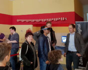 In einer Innenumgebung der Hochschule Coburg testen Menschen Virtual-Reality-Headsets. Eine Person in der Mitte trägt ein VR-Headset, während andere um sie herum zuschauen und interagieren. Im Hintergrund befindet sich eine VR-Demostation mit Informationsbannern.